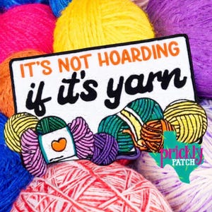 May include: A rectangular patch with the text "IT'S NOT HOARDING if it's yarn" in black and orange. The patch features illustrations of yarn balls in various colors, including pink, yellow, and green, set against a backdrop of colorful yarn.