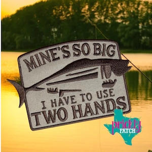 May include: Embroidered patch with the text "MINE'S SO BIG I HAVE TO USE TWO HANDS" and a fish design. The patch is gray with black stitching and a black border. The background shows a lake and trees.