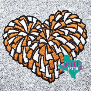 May include: An embroidered patch in the shape of a heart, composed of orange and white cheerleading pom-poms with a black stitched border. The design features a dense, textured pattern of overlapping strands, creating a full, rounded heart silhouette.