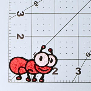 May include: A red embroidered patch in the shape of a cartoon ant with large eyes and antennae. The patch is on a white surface with black grid lines and numbers for measurement.