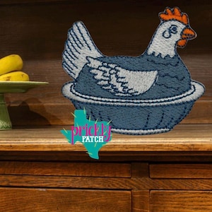 May include: Embroidered patch featuring a blue hen sitting in a nest. The hen has white and blue details, with an orange comb. The patch includes the text "prickly PATCH" in a turquoise and pink design.