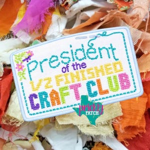 May include: A white rectangular patch with the words "President of the 1/2 Finished Craft Club" in colorful cross-stitch. The patch has a light blue border and floral accents. The background is a mix of colorful fabric scraps.