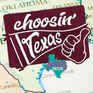 May include: A rectangular maroon embroidered patch featuring the white text "choosin' Texas" in a cursive script. The design includes a white pennant graphic and a white outline of a hand giving a thumbs-up gesture on the right side.