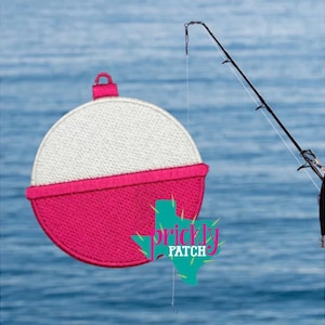 May include: A pink and white embroidered fishing bobber with a small loop at the top. A fishing rod is in the background. The bobber has the words "prickly patch" in a Texas-shaped graphic.