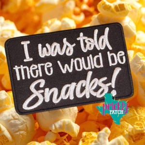 May include: A black rectangular patch with white text that reads "I was told there would be Snacks!" is set against a background of popcorn. The patch also features a colorful logo of a Texas outline with the words "Prickly Patch".
