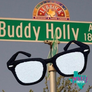 May include: An embroidered patch shaped like black-rimmed glasses with white lenses, positioned in front of a green street sign that reads Buddy Holly and a semi-circular sign above it featuring the text Legendary Lubbock Discover A Texas Legend.