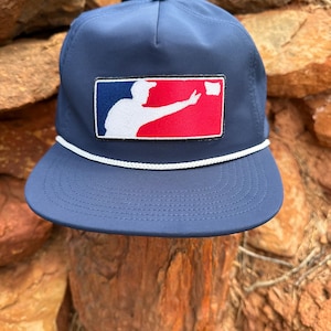 May include: A navy blue baseball cap featuring a rectangular patch on the front. The patch displays a white silhouette of a person throwing a white object against a blue and red background. A white braided cord runs across the base of the crown.