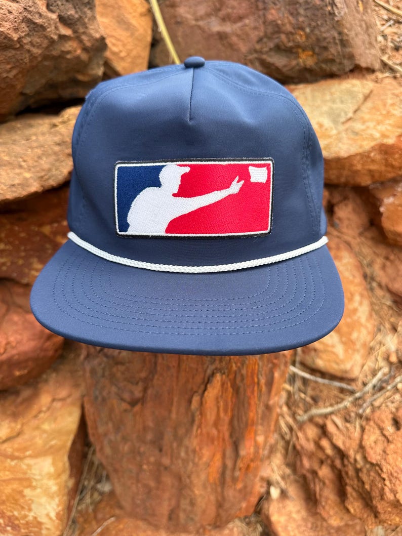 May include: A navy blue baseball cap featuring a rectangular patch on the front. The patch displays a white silhouette of a person throwing a white object against a blue and red background. A white braided cord runs across the base of the crown.