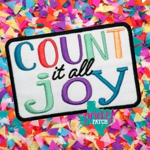 May include: A white rectangular patch with the words "COUNT it all JOY" embroidered in colorful letters. The patch has a black border and is set against a background of colorful confetti.