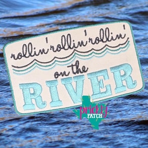 May include: Embroidered patch with the text "rollin' rollin' rollin' on the RIVER" in black and light blue. The patch has a rectangular shape with rounded corners and a light beige background. The Prickly Patch logo is at the bottom.