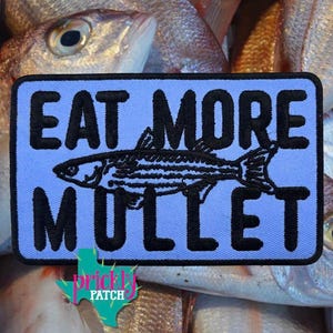May include: A rectangular embroidered patch with a light blue background and a black border. The text EAT MORE MULLET is stitched in black capital letters, with a black line drawing of a mullet fish centered between the two lines of text.