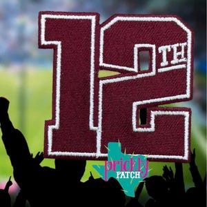 Può includere: Una toppa ricamata color bordeaux raffigurante il numero 12 con le lettere TH in un carattere più piccolo sulla destra. Il design presenta un bordo spesso cucito in bianco che delinea l'intera forma. La toppa è mostrata su uno sfondo sfocato di uno stadio.