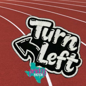 May include: Embroidered patch with the words "Turn Left" in white, outlined in black, with an arrow pointing left. The patch has a black background and a white border. The patch is on a red track surface.