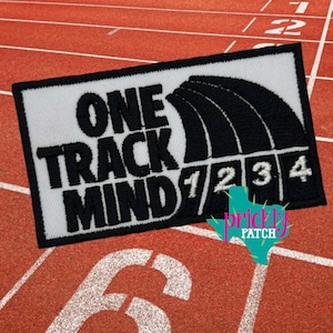May include: A rectangular patch with the words "ONE TRACK MIND" in black block letters on a white background. Below the text are black curved lines and numbers 1, 2, 3, and 4. The patch is on a red track surface.