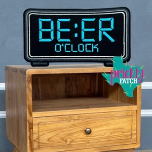 May include: A black digital clock with the words "BE:ER O'CLOCK" in bright blue. The clock sits atop a wooden nightstand with a drawer and open shelf. The clock has a black frame with a white border.