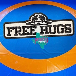 May include: A rectangular patch with the words "FREE HUGS" in black block letters on a white background. Above the text is a black graphic of two figures wrestling. The patch is set against a blue and orange background.