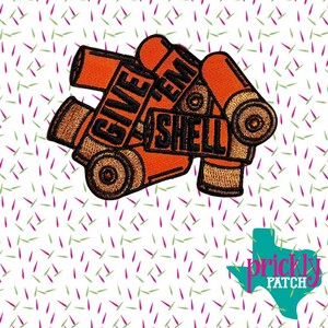 Könnte beinhalten: Gestickter Aufnäher mit orangefarbenen und braunen Schrotpatronen und dem schwarzen Schriftzug "GIVE 'EM SHELL". Der Aufnäher hat ein detailliertes Design und ist wahrscheinlich für das Aufnähen auf Kleidung oder Accessoires gedacht.