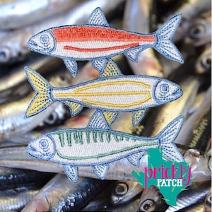 May include: Three embroidered fish patches in red, yellow, and green, each with a white body and blue accents. The patches are in a stack, with a blurred background of other fish. The patches are from Prickly Patch.