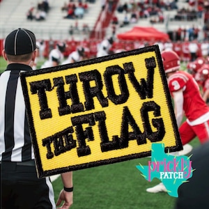 May include: A rectangular yellow embroidered patch with a black border and the black stitched text THROW THE FLAG. The patch is positioned over a blurred background of a football field with a referee and players in red uniforms visible.