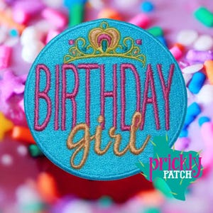 May include: A round, turquoise patch with the words "BIRTHDAY girl" embroidered in pink and gold. A gold and pink crown sits above the word "BIRTHDAY". The patch has a decorative border and a green and pink logo in the bottom right corner.