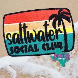 May include: A rectangular embroidered patch featuring a sunset gradient of red, orange, yellow, and teal stripes. The black text reads saltwater SOCIAL CLUB, accompanied by a black palm tree silhouette on the right side of the patch.