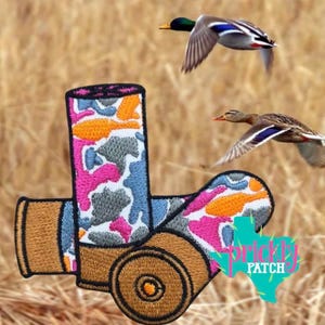 May include: An embroidered patch shaped like a shotgun shell with a camouflage pattern in shades of pink, orange, blue, and grey. The base and top of the shell are tan. Two mallard ducks are flying in the background above the patch.