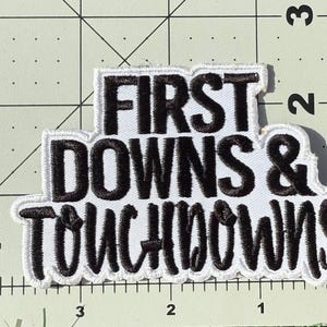 Puede incluir: Un parche bordado con fondo blanco y hilo negro que presenta el texto FIRST DOWNS & TOUCHDOWNS. El parche est&aacute; colocado sobre una base de corte con l&iacute;neas de cuadr&iacute;cula y n&uacute;meros visibles, mostrando que mide aproximadamente 8,9 cm de ancho.