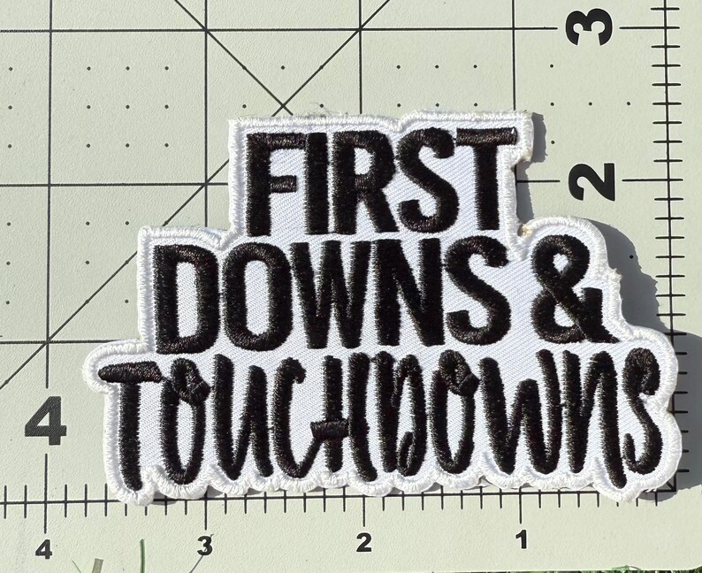 Puede incluir: Un parche bordado con fondo blanco y hilo negro que presenta el texto FIRST DOWNS & TOUCHDOWNS. El parche est&aacute; colocado sobre una base de corte con l&iacute;neas de cuadr&iacute;cula y n&uacute;meros visibles, mostrando que mide aproximadamente 8,9 cm de ancho.