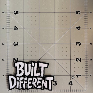 May include: A white and black embroidered patch with the text "BUILT DIFFERENT" in a bold, stylized font. The patch is positioned on a cutting mat with imperial markings, showing the patch is approximately 3.5 inches wide and 2 inches tall.