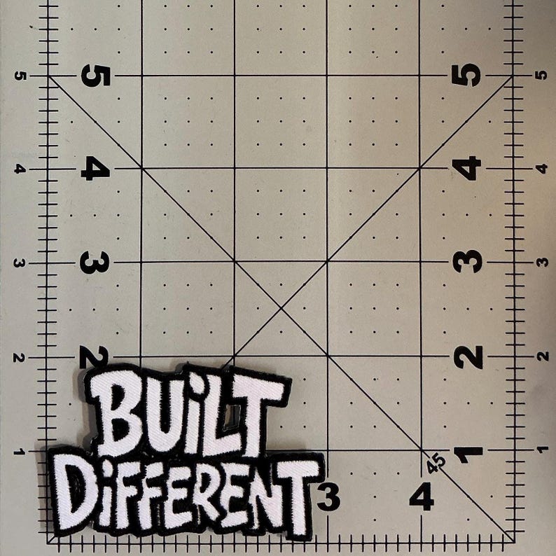 May include: A white and black embroidered patch with the text "BUILT DIFFERENT" in a bold, stylized font. The patch is positioned on a cutting mat with imperial markings, showing the patch is approximately 3.5 inches wide and 2 inches tall.