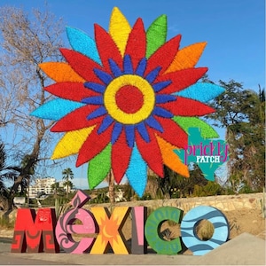 May include: A vibrant, multi-colored floral sculpture with red, orange, yellow, green, and blue petals. The word "MÉXICO" is displayed in large, colorful letters in front of the sculpture, with a blue sky background.