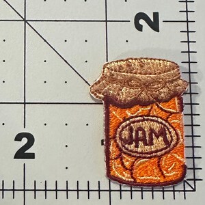 Peut inclure: &Eacute;cusson brod&eacute; repr&eacute;sentant un pot de confiture. L'&eacute;cusson montre un pot orange avec un couvercle marron et une &eacute;tiquette indiquant "JAM". L'&eacute;cusson est pos&eacute; sur un fond blanc avec des lignes de quadrillage noires.