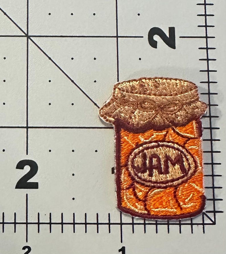 Peut inclure: &Eacute;cusson brod&eacute; repr&eacute;sentant un pot de confiture. L'&eacute;cusson montre un pot orange avec un couvercle marron et une &eacute;tiquette indiquant "JAM". L'&eacute;cusson est pos&eacute; sur un fond blanc avec des lignes de quadrillage noires.