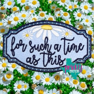 May include: Embroidered patch with the phrase "for such a time as this" in black script on a light blue background. The patch features daisy flower designs and a dark blue border. The background is a field of daisies.