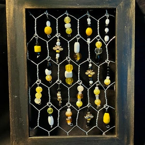 May include: A framed piece of art featuring a wire mesh background with various yellow, white, and black beads and charms hanging from it. The charms include small bees and other decorative elements. The frame is black with a distressed finish.