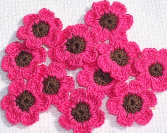 May include: A collection of handmade crochet flowers in a vibrant fuchsia pink with brown centers. The flowers are arranged on a white, textured surface, showcasing their detailed petal design. These decorative flowers are ideal for crafting projects.