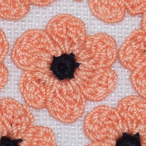 May include: Multiple crocheted flower appliques arranged on a white grid fabric. Each flower features five rounded peach-colored petals surrounding a solid black circular center. The yarn texture is visible throughout the floral pattern.