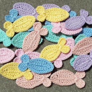 May include: A collection of colorful, crocheted fish in various pastel shades. The fish are in shades of yellow, pink, blue, green, and purple. Each fish has a three-lobed tail and a rounded body. The fish are scattered on a neutral surface.