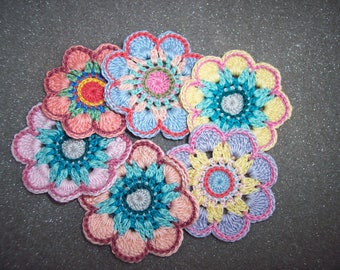 May include: A collection of six handmade crochet flowers in various pastel colors. Each flower features a central design with multiple layers of petals, showcasing a mix of pink, blue, yellow, and green hues. The flowers are arranged on a textured, gray surface.