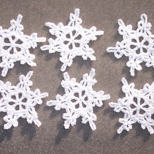 May include: Six white, crocheted snowflakes are arranged on a textured, gray surface. Each snowflake has a delicate, openwork design with six points radiating from a central circle. The snowflakes are evenly spaced, showcasing their intricate details.