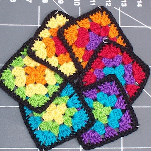 May include: A collection of colorful, square crochet pieces. Each piece features a floral design in vibrant hues of yellow, orange, red, purple, green, and blue, outlined with black yarn. The pieces are arranged in a layered pattern.