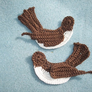 May include: Two bird-shaped crochet appliques featuring dark brown yarn for the head, back, and tail, contrasted with white yarn for the chest and belly. Each piece is crafted with visible stitch patterns, depicting a bird in a side profile view.