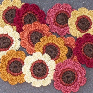 May include: A collection of handmade crochet flowers in various colors, including yellow, orange, red, and pink. Each flower has a dark brown center and is arranged on a textured gray surface. These decorative flowers are ideal for crafting projects.