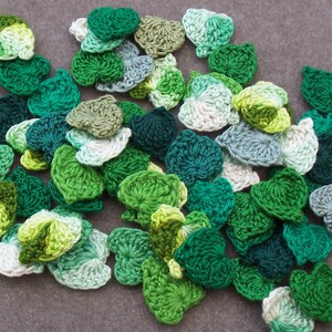 May include: A collection of handmade crochet leaf appliques in various shades of green, from light lime to deep forest. The leaves are scattered on a gray surface, showcasing their detailed texture and varied hues. Ideal for crafting and embellishments.