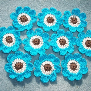 May include: A collection of ten crocheted flowers. Each flower has a bright turquoise blue petal design, a white inner ring, and a brown center. The flowers are arranged on a light blue surface.