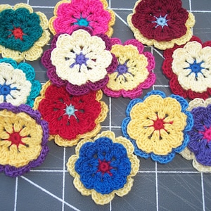 May include: A collection of colorful, crocheted flower appliques in various sizes. The flowers feature multiple layers of petals in shades of yellow, red, blue, purple, and pink, with contrasting center colors. The appliques are arranged on a dark surface.