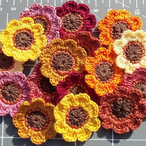 May include: A collection of crocheted flower appliques in various colors including yellow, orange, red, pink, and cream, each featuring a dark brown center. The flowers are arranged on a gray cutting mat with visible white grid lines and centimeter markings.