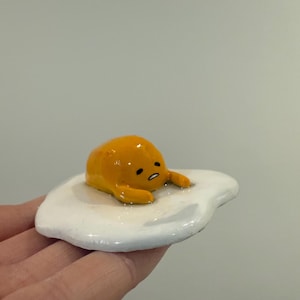 Puede incluir: Una figurita de cerámica amarilla y blanca de un personaje de dibujos animados con forma de huevo con una expresión triste. El personaje está acostado de lado sobre una base de cerámica blanca con forma de huevo.