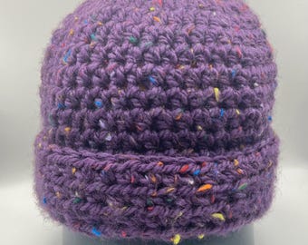 May include: A purple crocheted beanie hat with colorful flecks. The hat has a folded brim and a textured stitch pattern. The yarn appears to be a soft, warm material, suitable for cold weather.