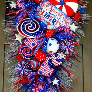 May include: A patriotic wreath with red, white, and blue decorations. It features star-spangled ribbons, spiral candies, and a sign that reads "Sweet Land of Liberty." The wreath is adorned with star-shaped ornaments and faux strawberries.